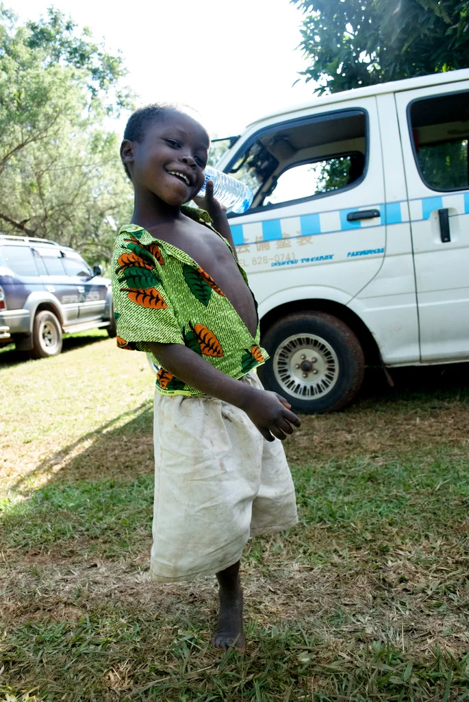 Smiling young boy holding a plastic water bottle, standing on grass outdoors near parked vehicles, wearing a colorful shirt and light-colored shorts in Uganda.