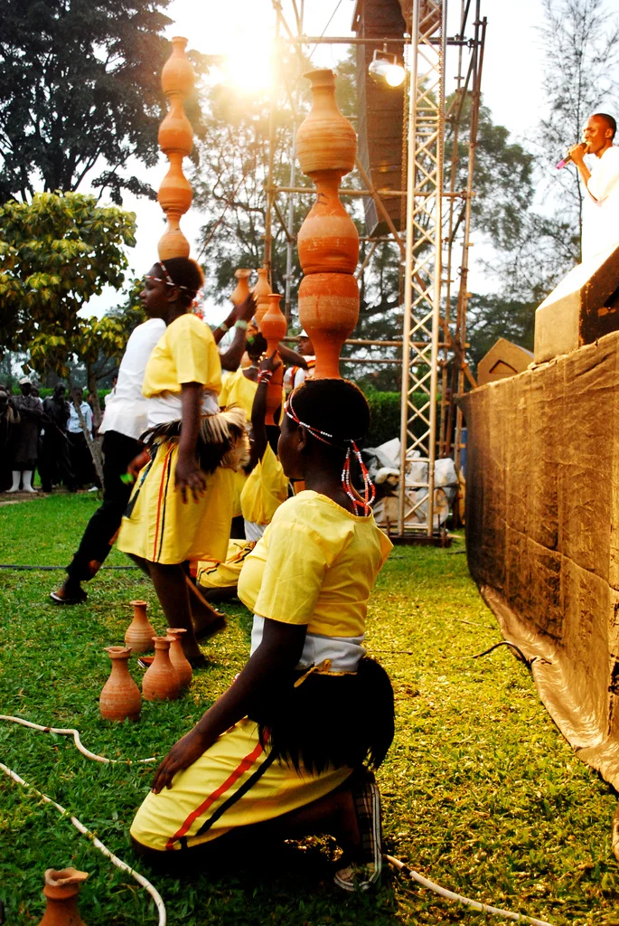 Children in yellow costumes performing a traditional dance with clay pots balanced on their heads at an outdoor event called Bayimba during sunset in Uganda.