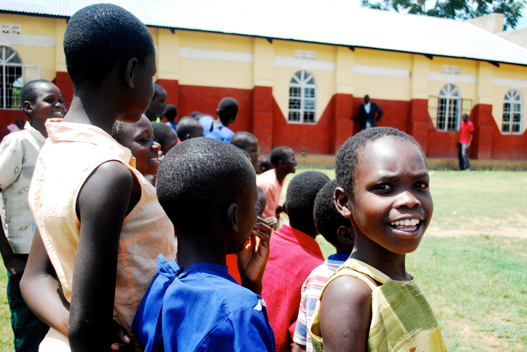 Group of children standing outside on a grassy area, some smiling and talking, with a building in the background in Uganda.