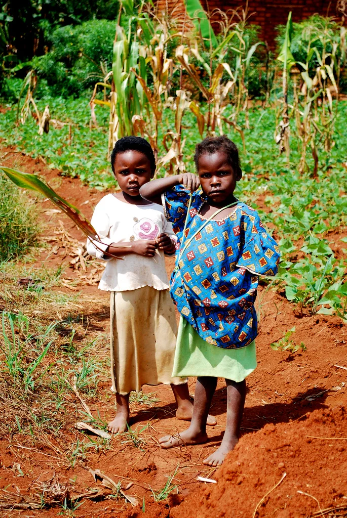 Two children standing barefoot in a rural garden with tall corn and green plants in the background, holding a leaf and posing for the camera in Uganda.