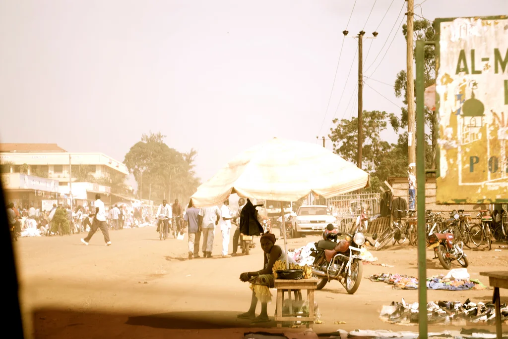 Street scene in a marketplace with people walking, motorcycles parked, and vendor stalls with umbrellas in a dusty environment in Uganda.