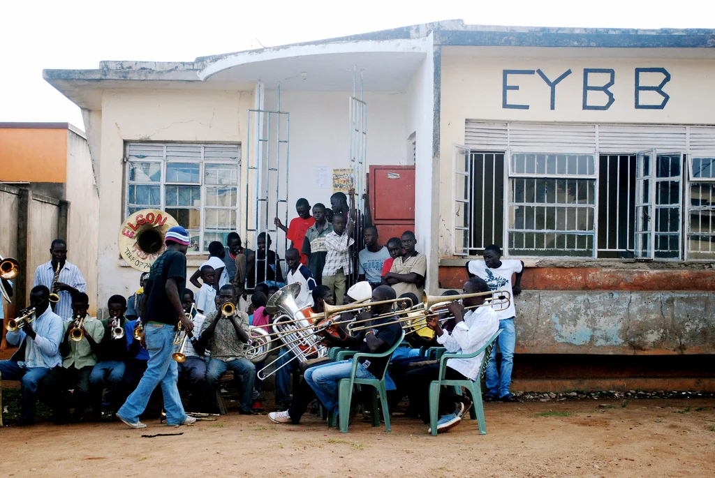 A group of children and teenagers playing musical instruments outside a building with the letters EYBB on it, likely a school or community center in Uganda.