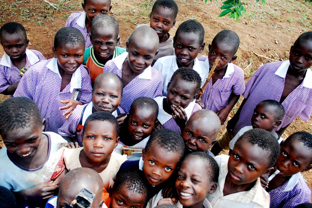 Group of young children outdoors, most wearing purple and white checkered school uniforms, some smiling and looking at the camera in Uganda.