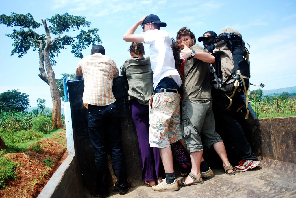 A group of six people on a narrow bridge or walkway outdoors, with greenery and trees in the background, all facing away or partially turned away from the camera in Uganda.