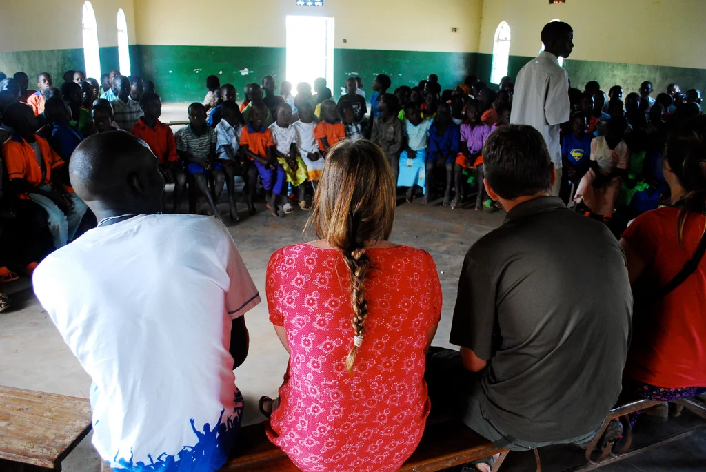 Group of children and adults seated inside a classroom with green and yellow walls, listening to a speaker standing at the front in Uganda.
