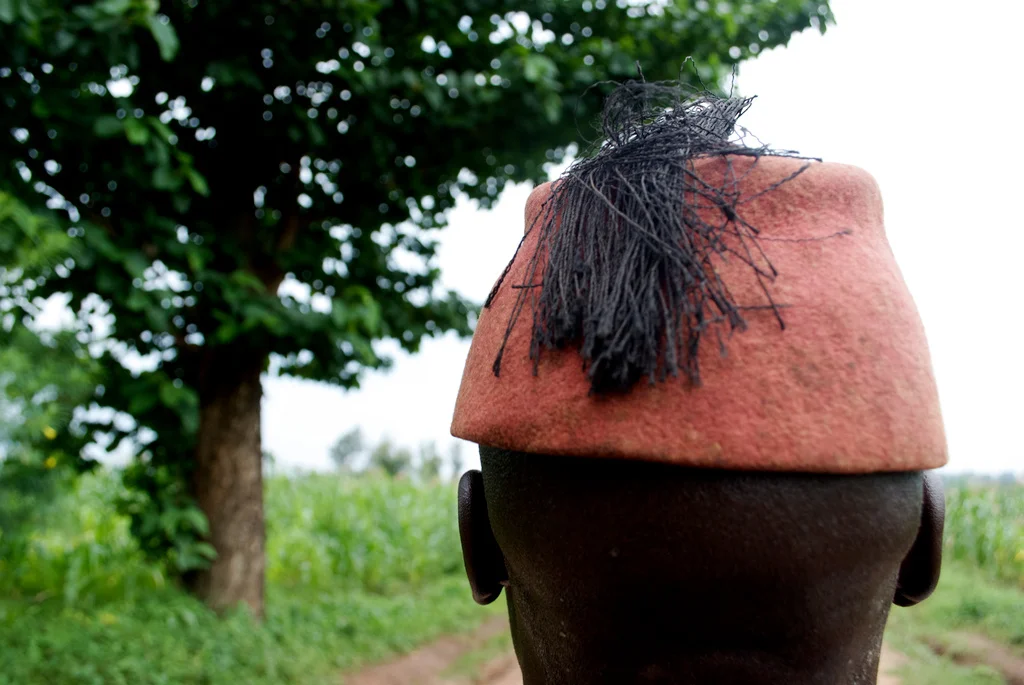 A person wearing a red felt hat with black hair on top, standing outdoors with green trees and plants in the background in Uganda.