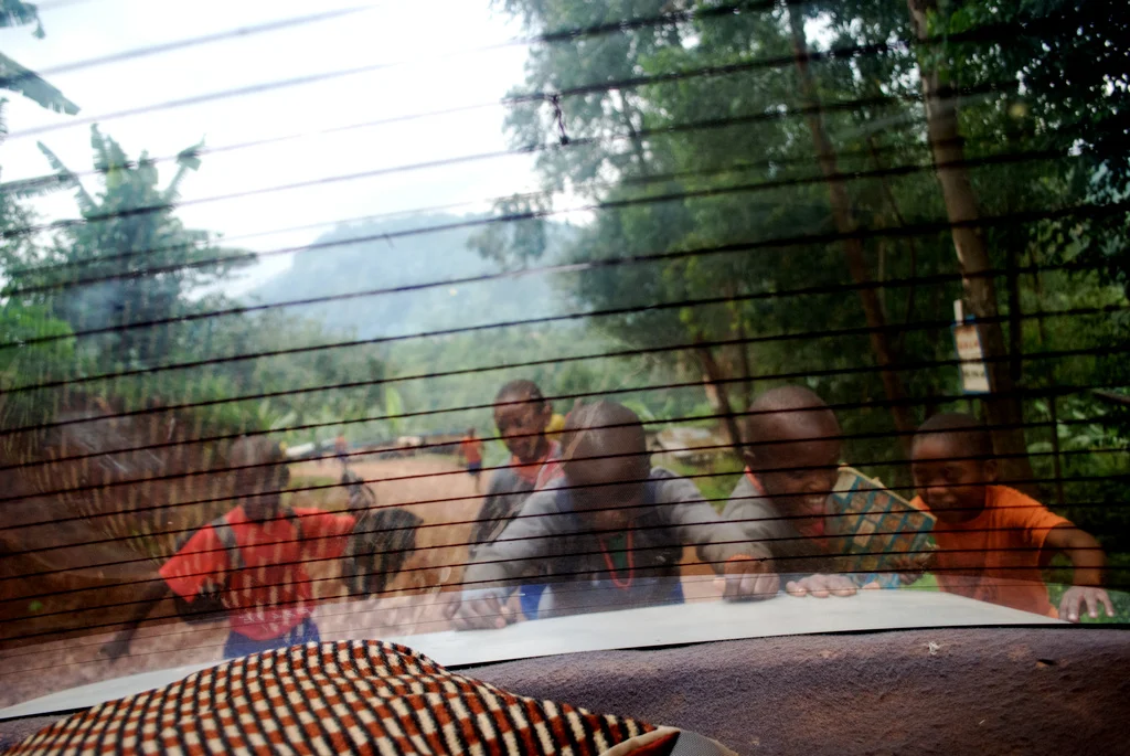 Children looking through a car window at camera with trees and dirt road in background in Uganda.