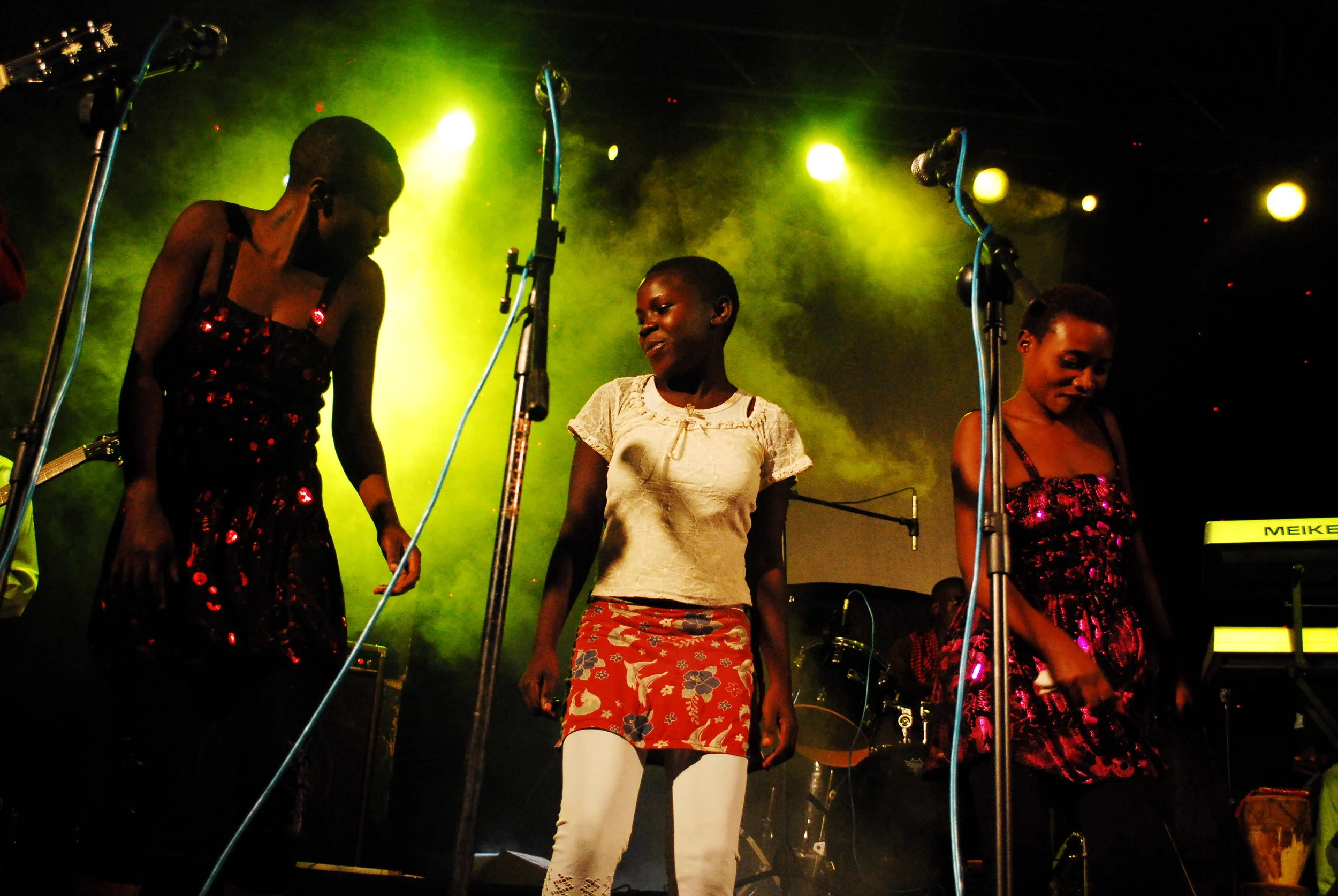 Three women on stage with musical instruments, illuminated by green and yellow stage lights, in a live performance setting at Bayimba in Uganda.