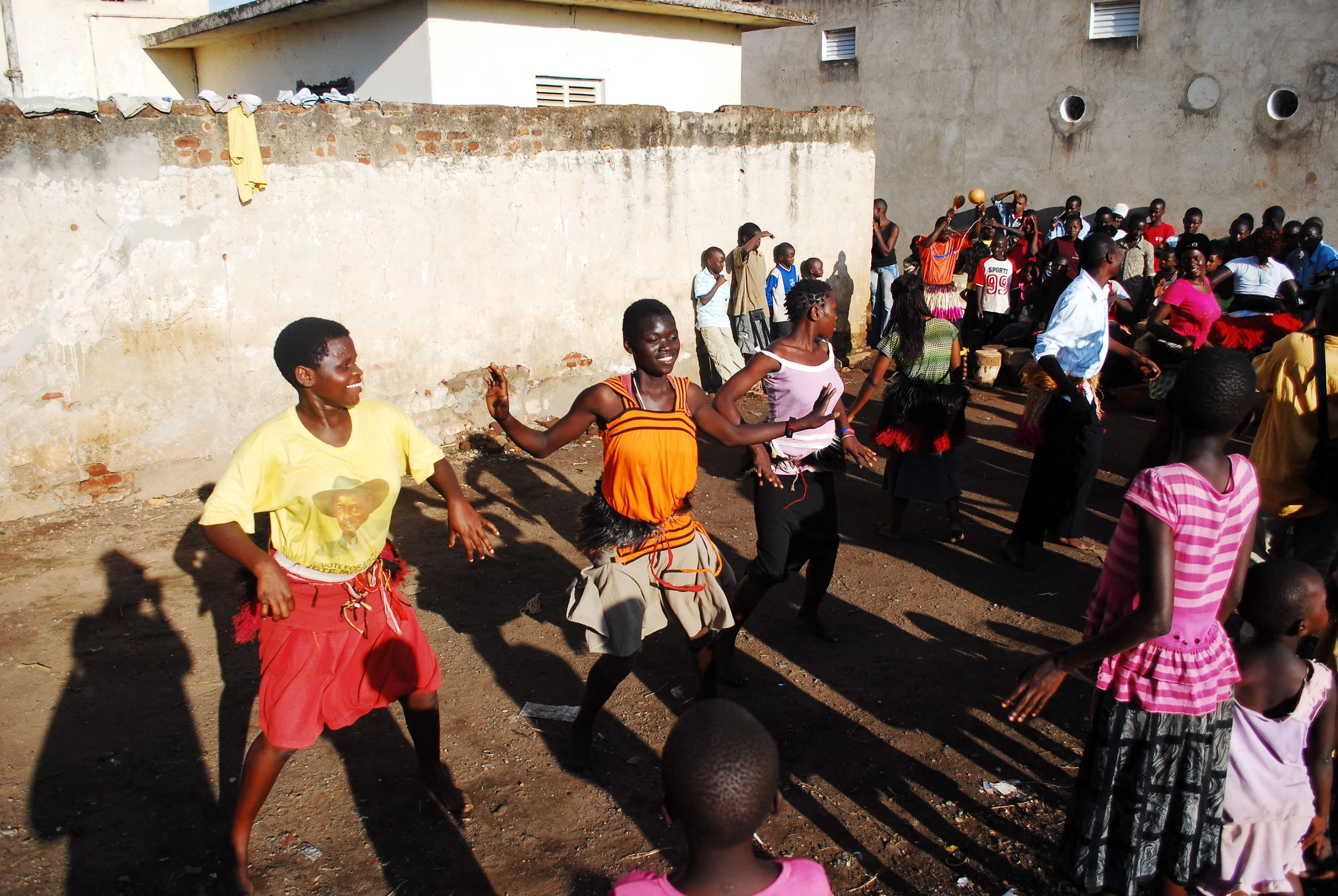 People dancing and socializing outdoors in a dirt area with cold concrete walls and a group of children and adults in Uganda. The scene appears lively and festive.
