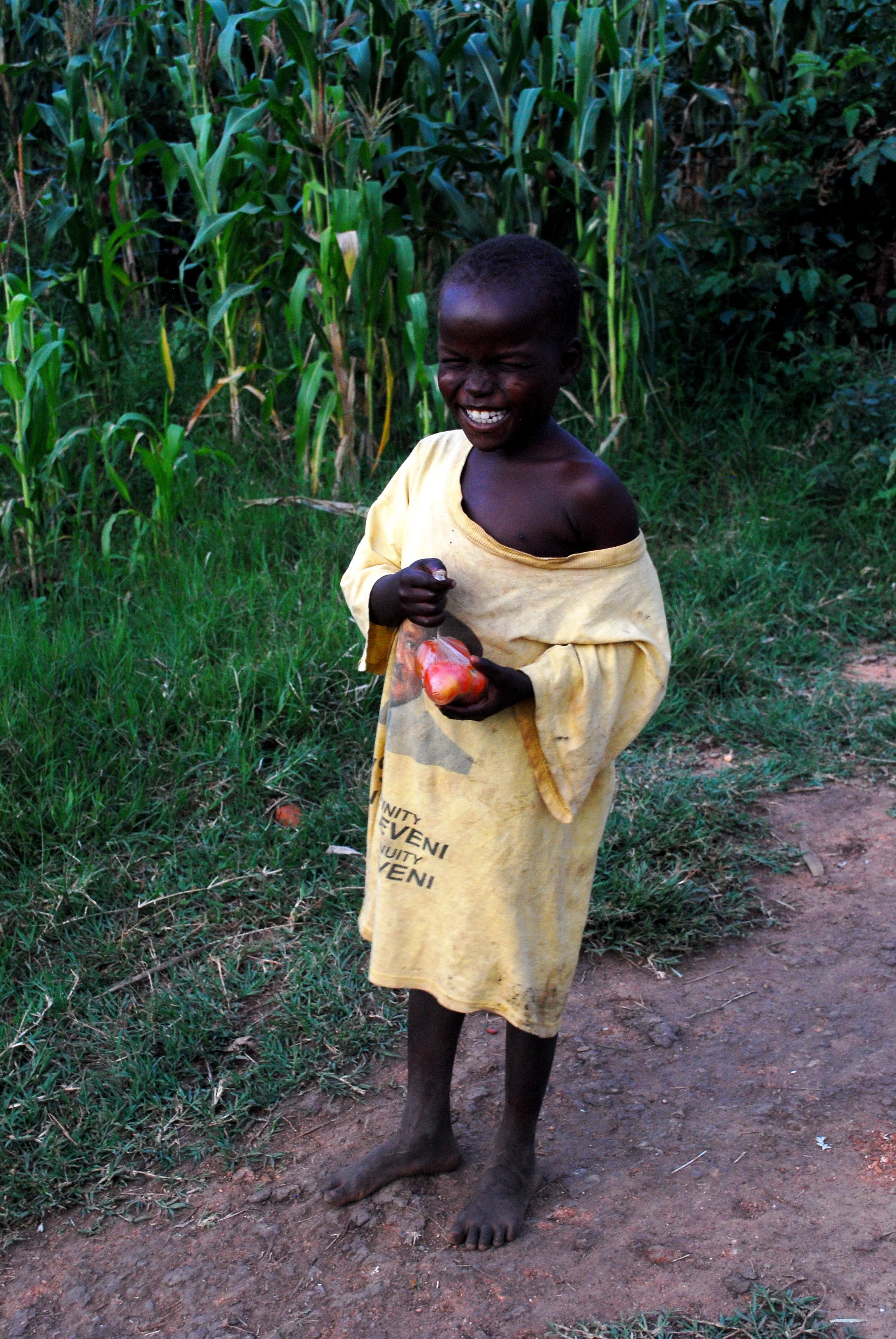A young boy smiling and holding a bag of tomatoes, standing barefoot on a dirt path near a cornfield in Uganda.