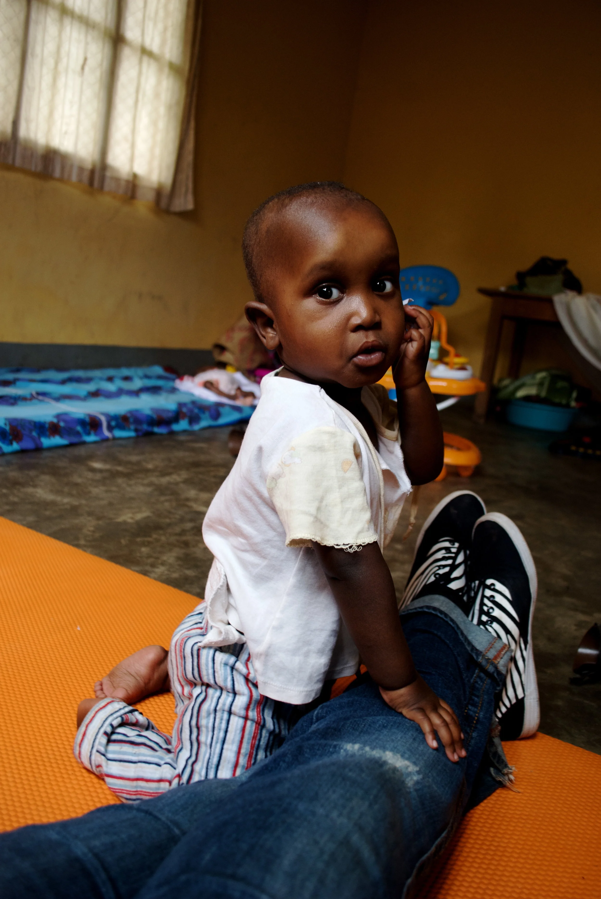 A young child with short hair sitting on an orange mat, looking at the camera, with a hand on their cheek, in a room with yellow walls and a window. There are toys and bedding in the background in Uganda.