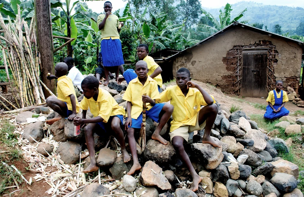 Group of children in yellow shirts and blue skirts or shorts, some sitting and some standing, outdoors on a rocky area with a rustic house and lush green banana plants in the background in Uganda.