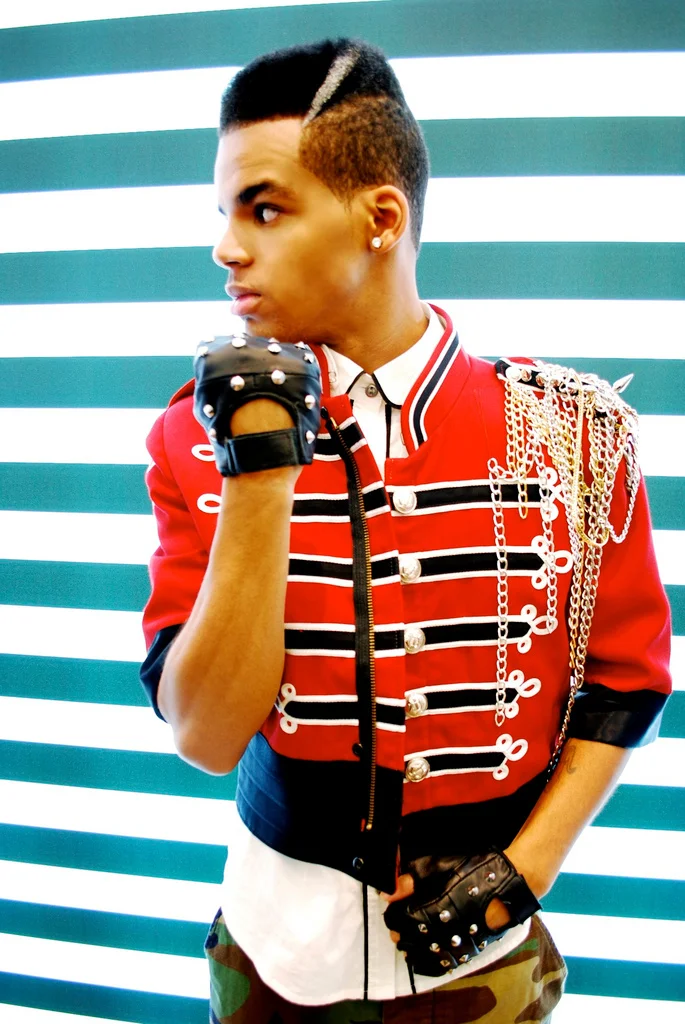 Young man with a mohawk hairstyle wearing a red military-style jacket with gold chains and epaulets, black and white gloves, in front of striped background