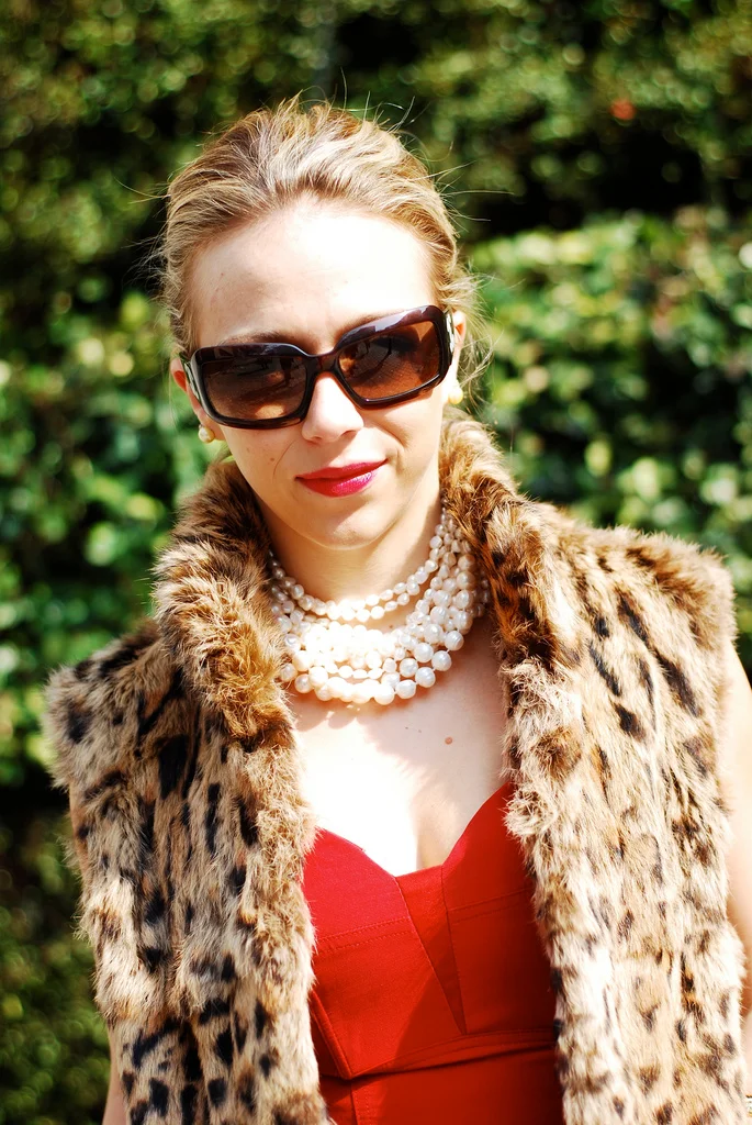A woman wearing large black sunglasses, red lipstick, a pearl necklace, a red dress, and a leopard print faux fur vest outdoors with greenery background.