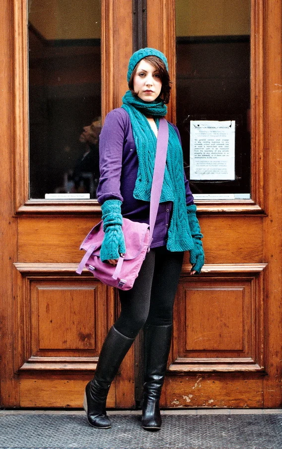 Young woman standing in front of wooden door, wearing a teal knit hat, scarf, gloves, purple jacket, black leggings, and knee-high black boots, carrying a pink shoulder bag.