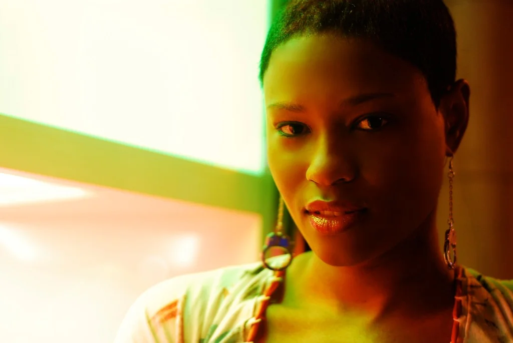 Close-up of a woman with dark skin, short hair, and jewelry, looking at the camera with a colorful illuminated background.