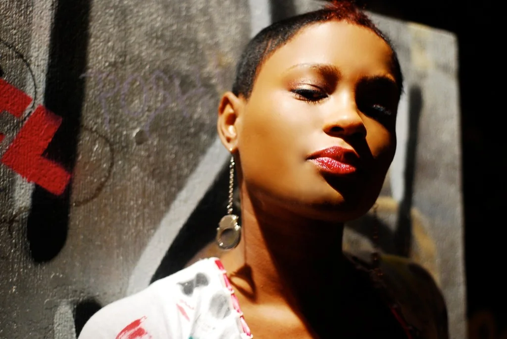 A woman with short hair and earrings leaning against a graffiti-covered wall.