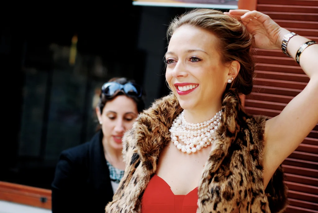 Two women smiling outdoors, one in a red dress with pearl necklaces and a fur vest, the other in black with goggles on her head.