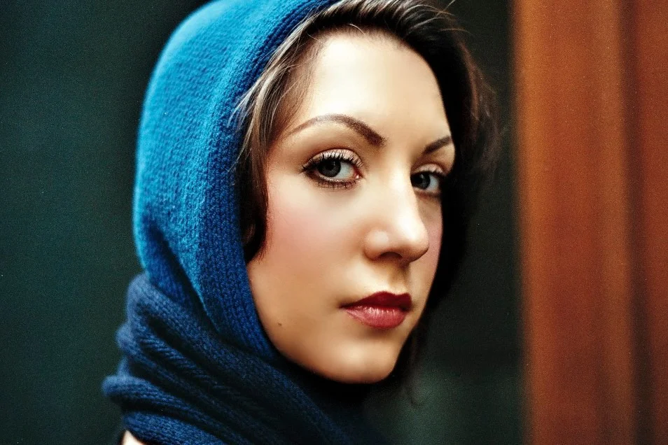 A close-up portrait of a woman wearing a blue headscarf, with makeup and piercing eyes, looking at the camera.
