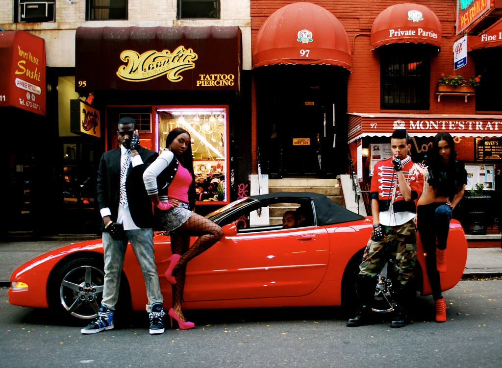 Group of four young people posing in front of a red sports car on an urban street, with storefronts and signs in the background.