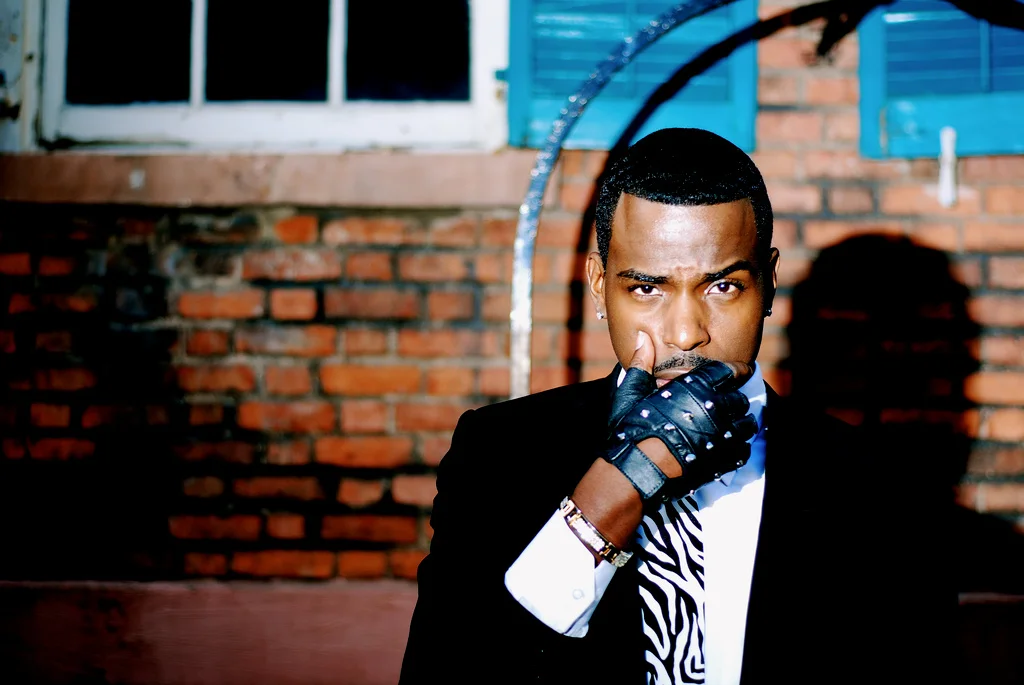 Young man wearing a black suit, striped tie, leather gloves, standing in front of a brick wall, with a window above.
