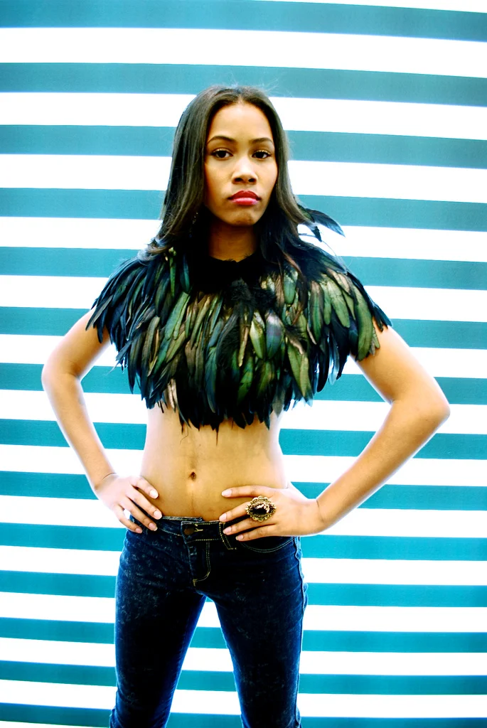 A young woman with medium skin tone and straight dark hair poses with hands on hips against a blue and white striped background. She wears a feathered Top with dark green and black feathers, dark jeans, and a large decorative ring on her right hand.