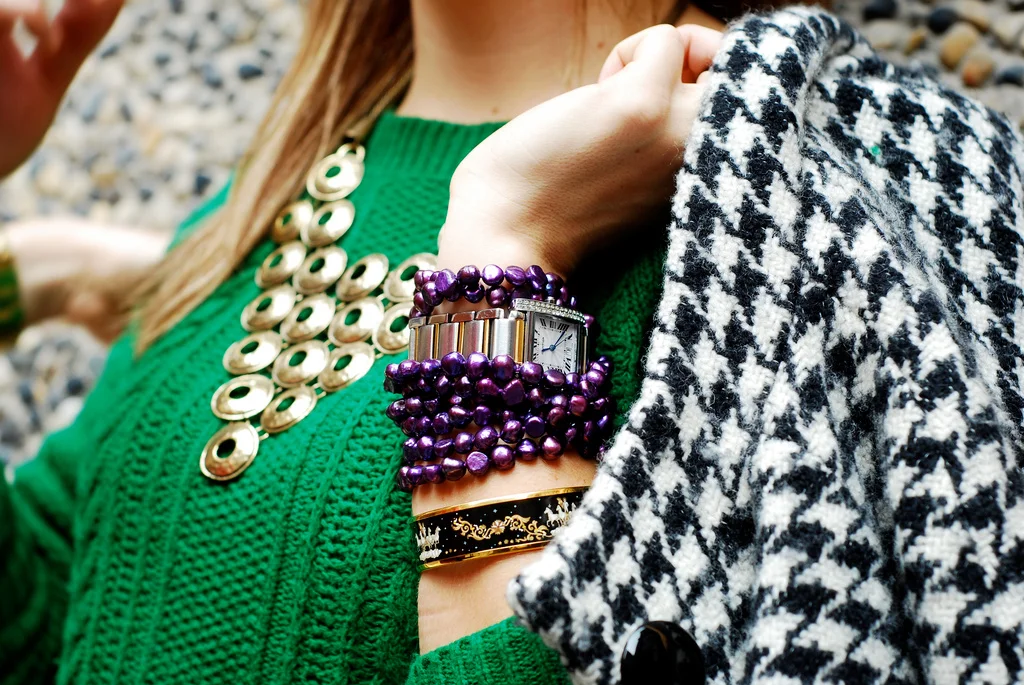 Close-up of a woman wearing a green sweater with layered jewelry including a large pearl necklace, a gold and purple beaded bracelet, a black and gold bracelet, and a silver watch, holding a black and white houndstooth patterned scarf.