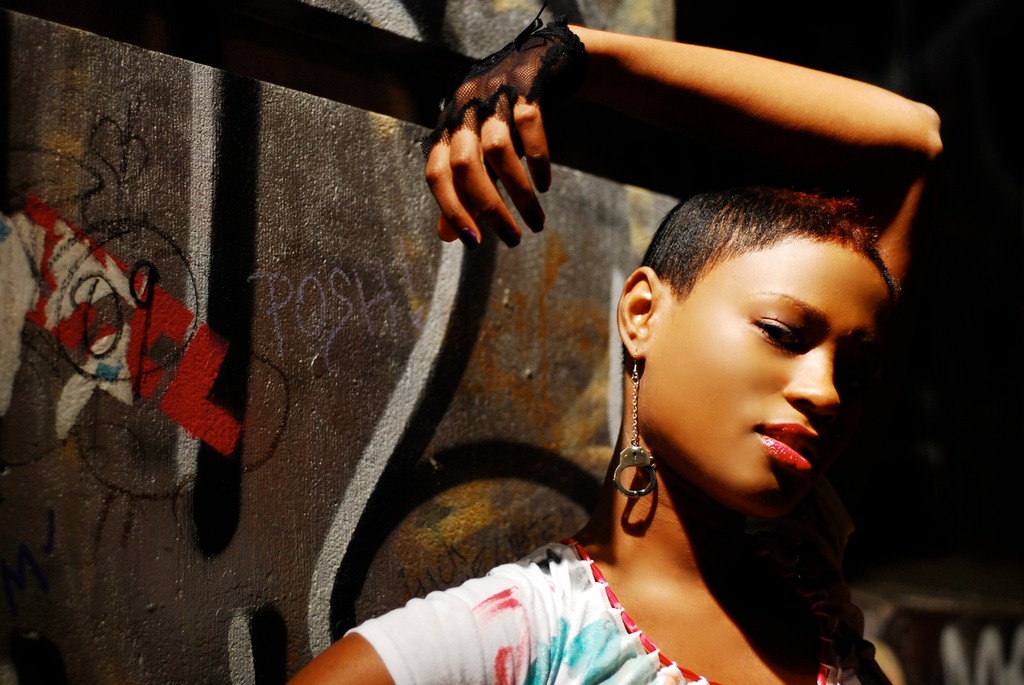 A woman with short hair poses against a graffitied wall, wearing an earring shaped like a hand and a fingerless glove, with her arm resting on the wall.