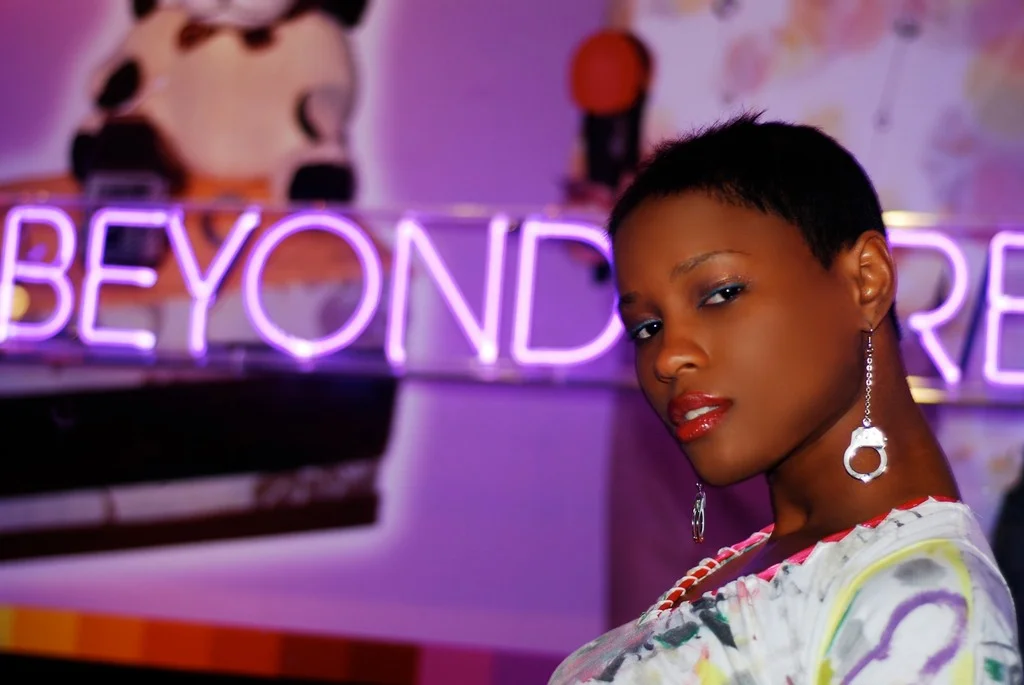 A woman with short black hair and red lipstick posing in front of a neon sign that says 'BEYOND'. She is wearing earrings and a colorful, patterned top, with a confident expression.