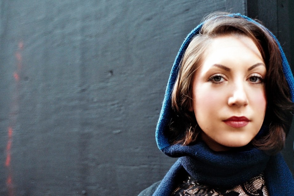 Close-up of a young woman with dark brown hair, wearing a dark blue hooded scarf, standing against a black wall.