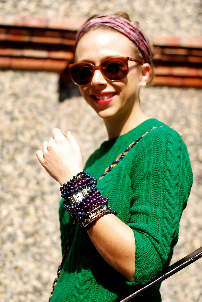 A woman wearing a pink headband and sunglasses, dressed in a green sweater, smiling outdoors with a gravel background and brick wall behind her, accessorized with multiple bracelets.