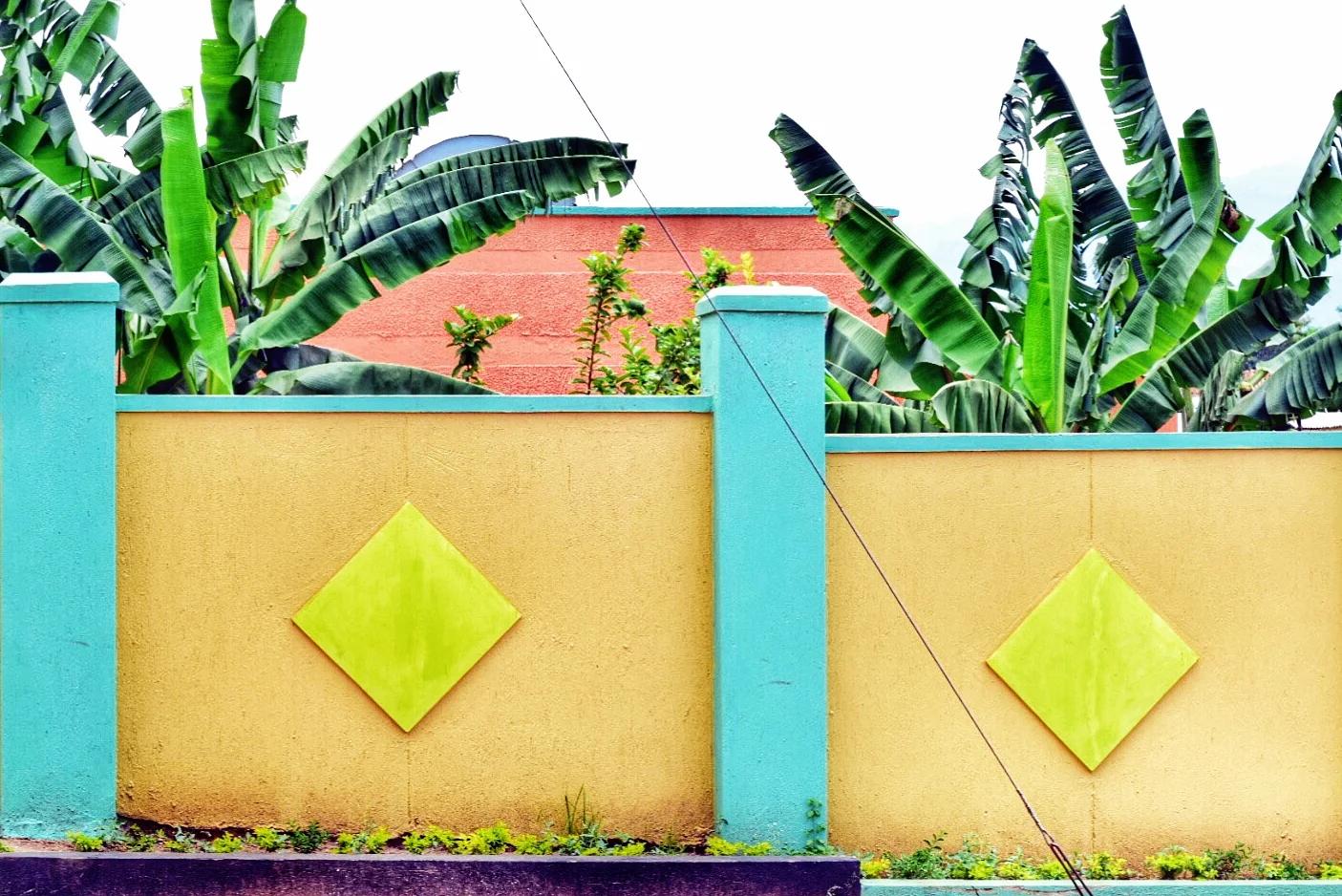 Colorful exterior wall with blue columns, yellow main wall, and green diamond-shaped accents, with banana trees behind in Mbale, Uganda.
