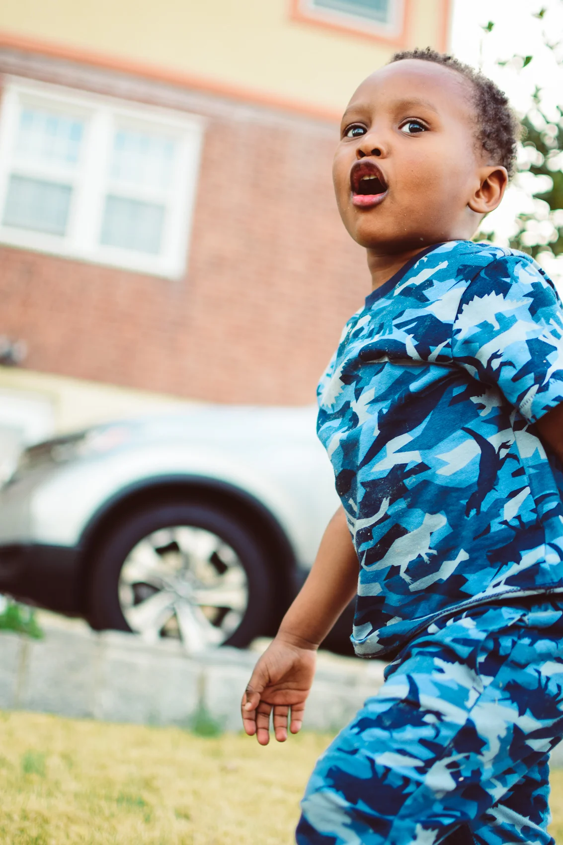 A young boy wearing a blue camouflage shirt standing outside on the grass near a brick house and a car.