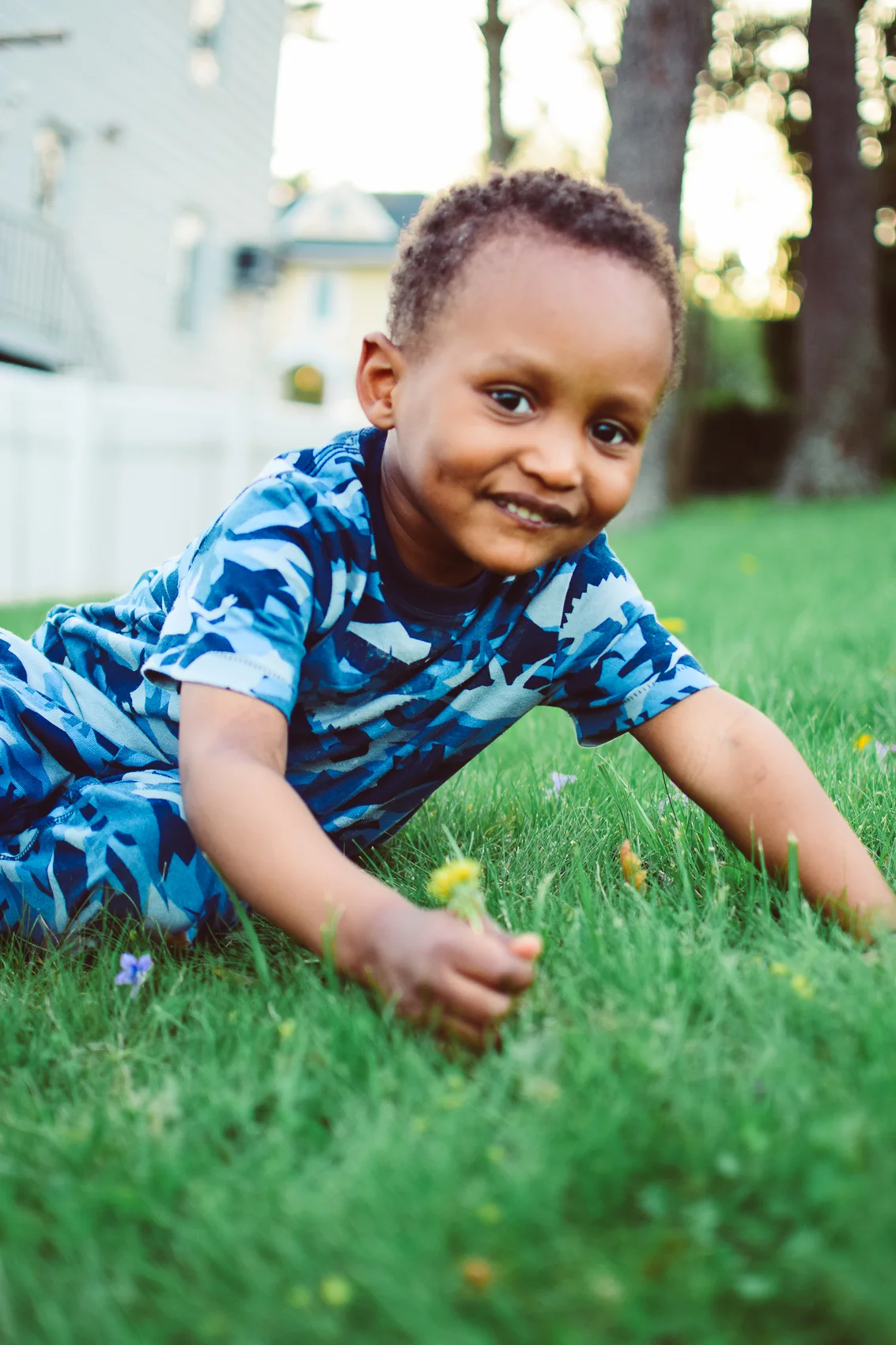 A young boy wearing a blue camouflage shirt is crawling on green grass in a backyard, smiling at the camera with trees and houses in the background.