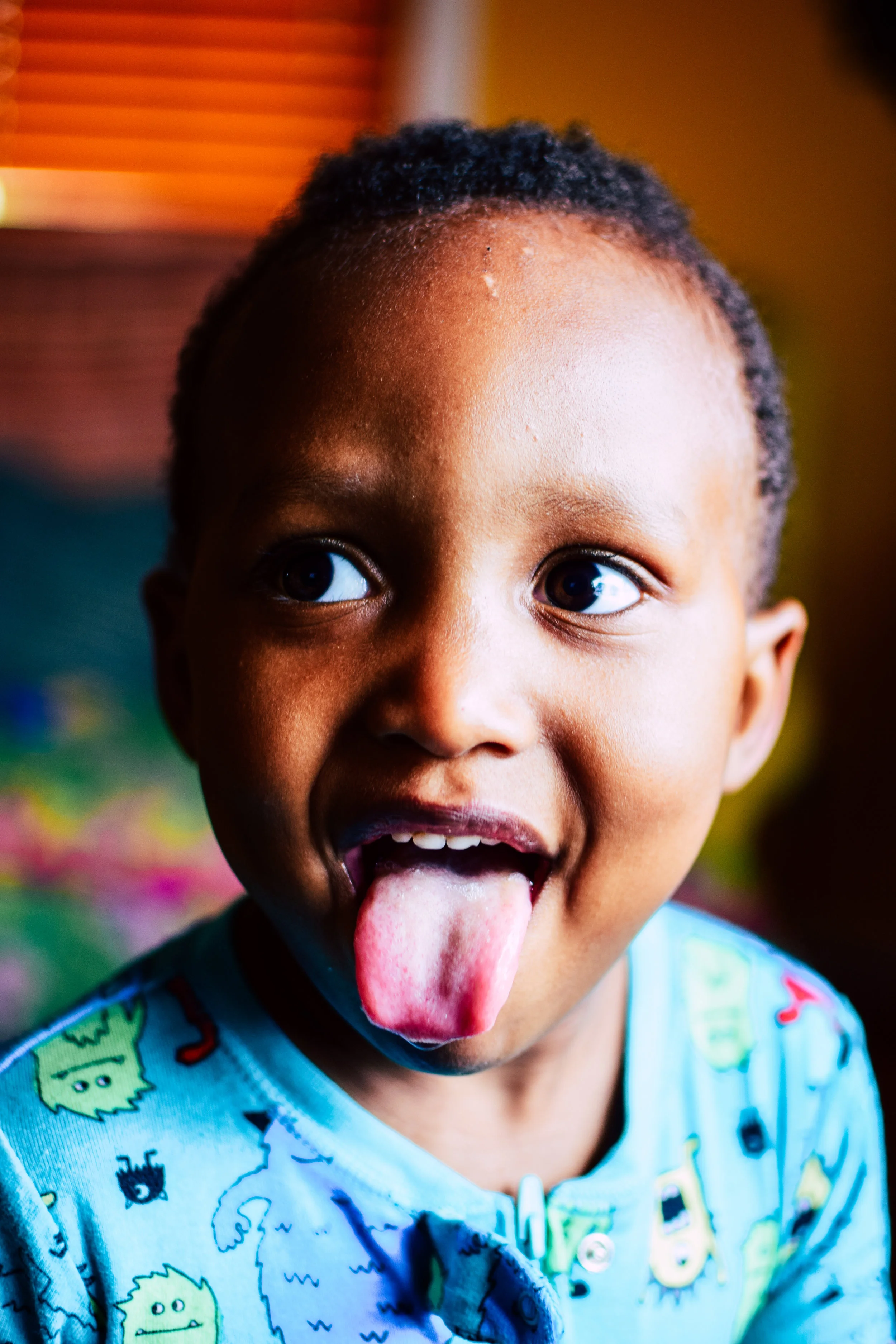 Close-up of a young child with short curly hair, sticking out tongue, wearing a light blue shirt with colorful cartoon patterns, indoors with warm lighting.
