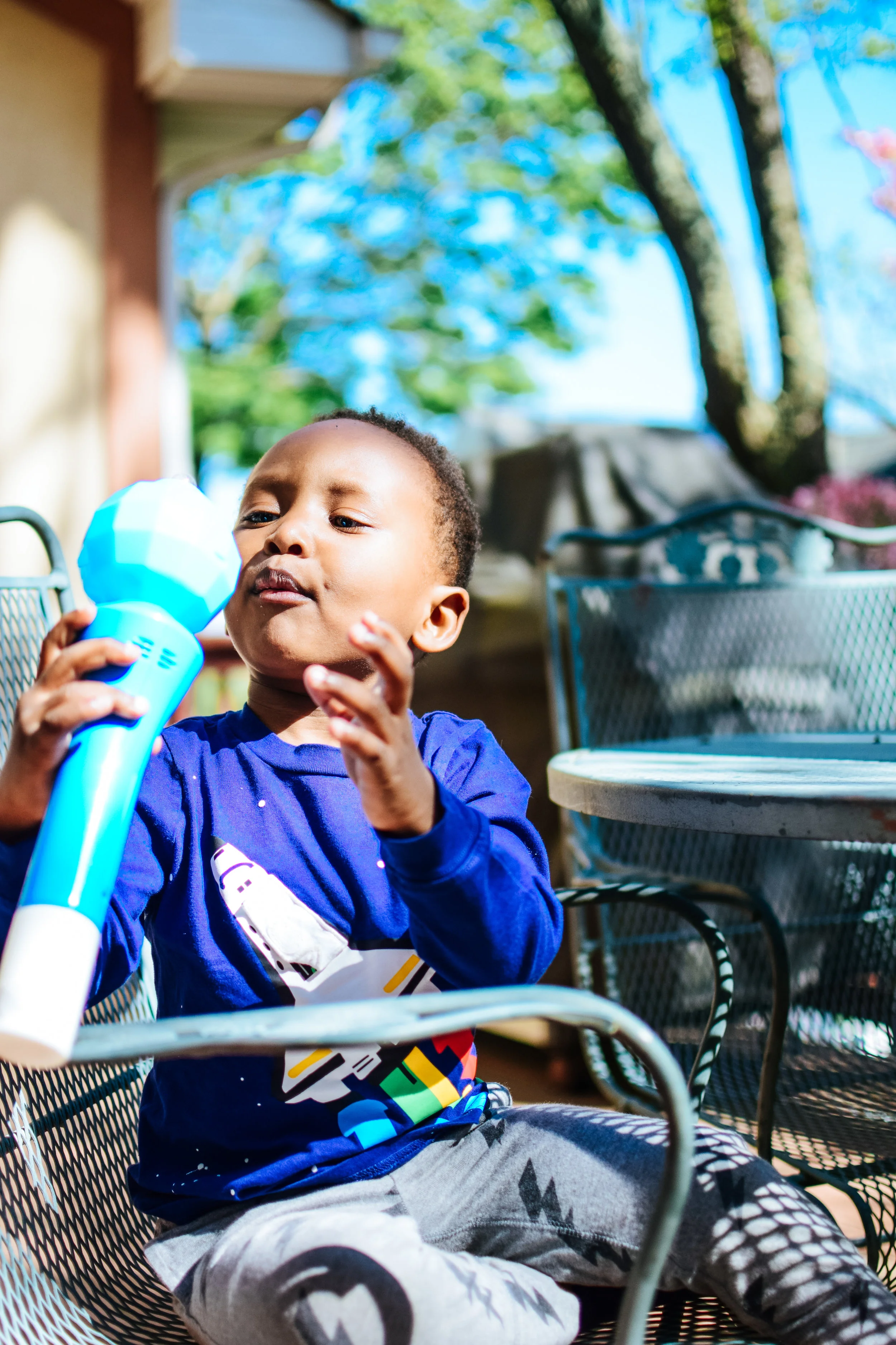 Young boy in a blue long sleeve shirt playing with a blue and white toy while sitting outside on a sunny day.