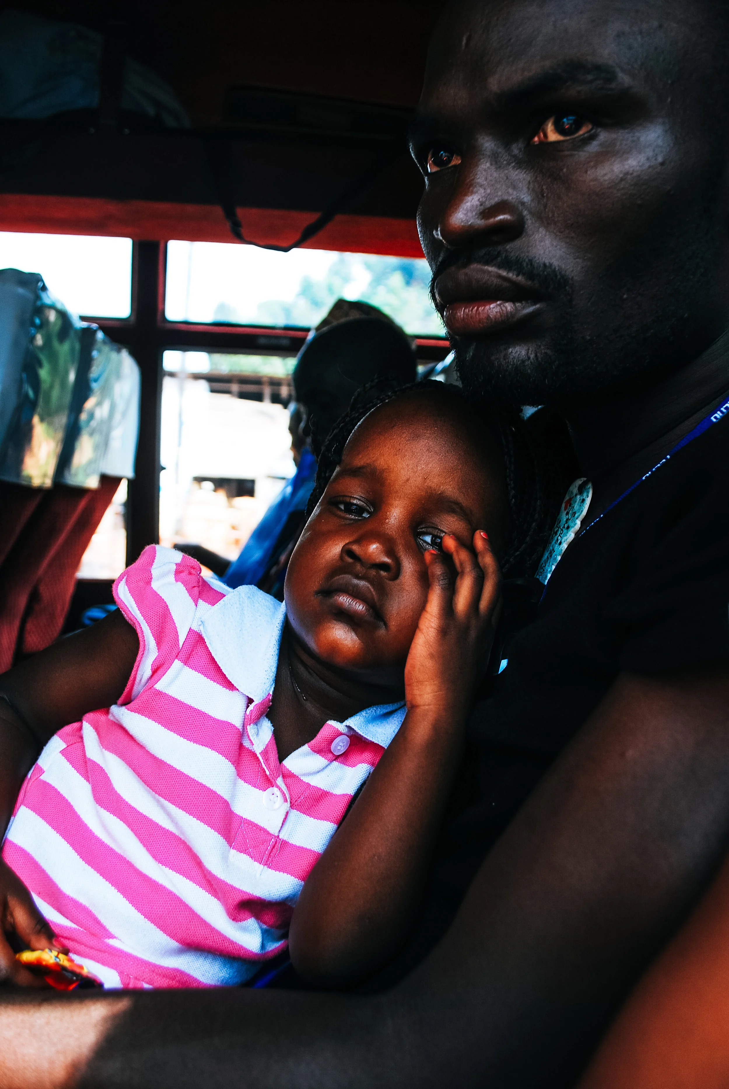 A young girl with a tired or sad expression, resting her head against an older man's chest, sitting inside a vehicle with other passengers visible in the background in Uganda, East Africa.