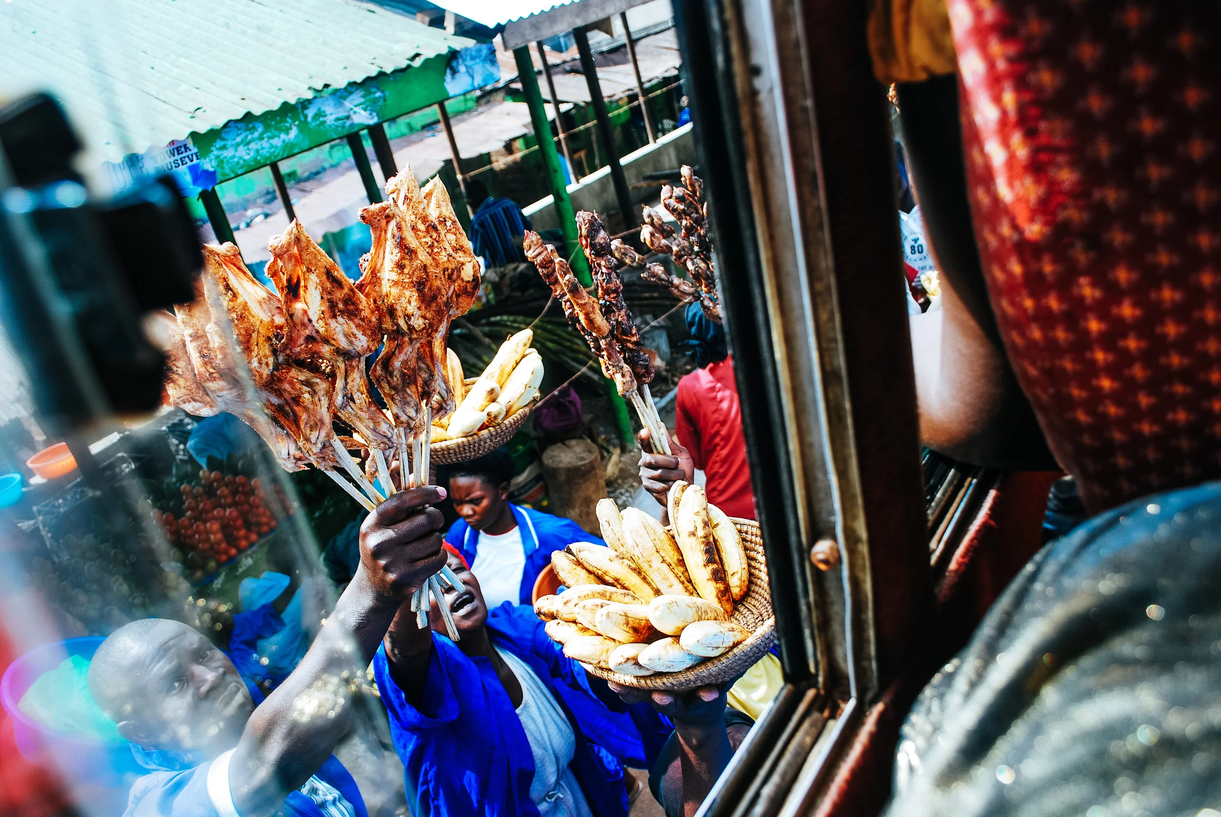 Street vendors selling grilled meat and bananas, viewed through a shop window, with people in the background in a busy market area in Uganda, East Africa.