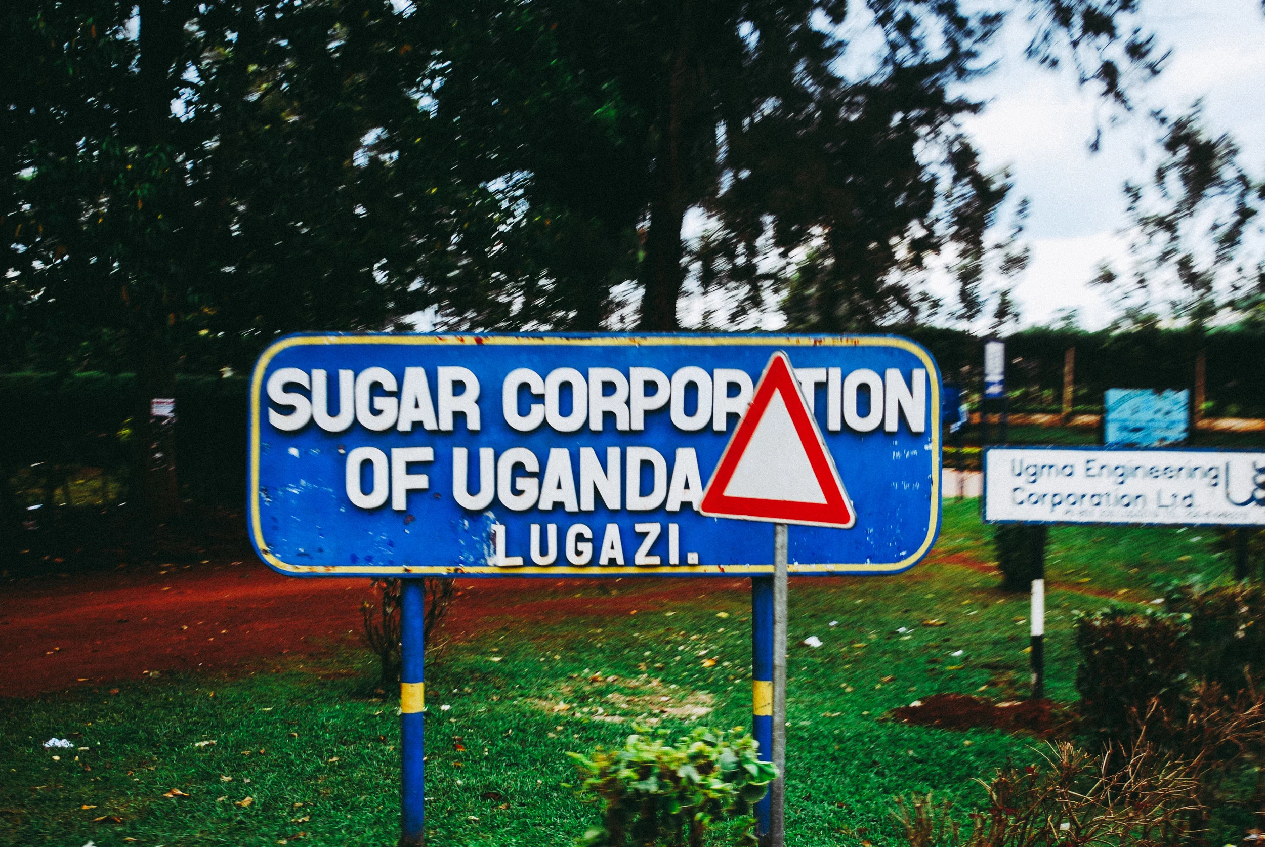 Blue sign with white text reads 'SUGAR CORPORATION OF UGANDA LUAGZI' with a red and white triangular warning sign on it, placed outdoors with grass and trees in the background.