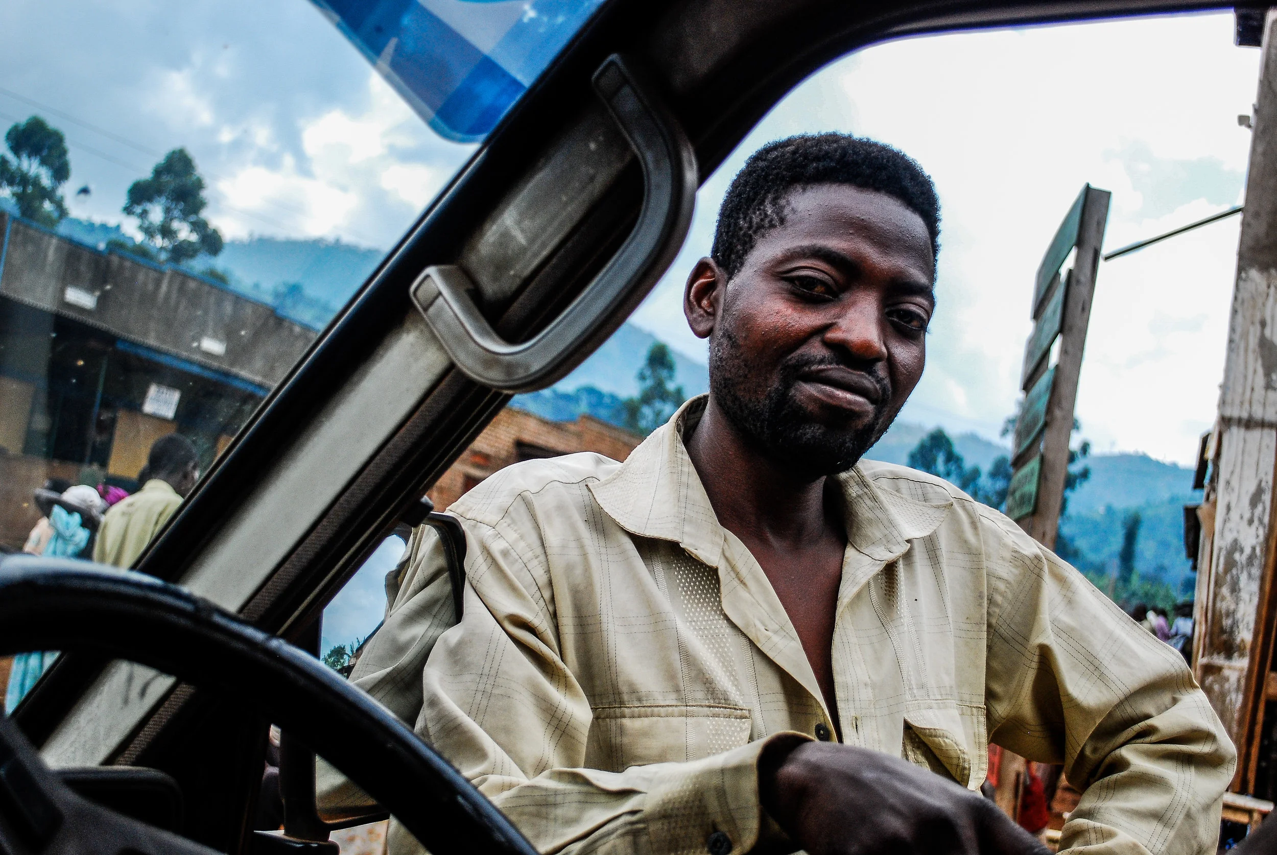 A man leaning on a vehicle engaged in conversation, with mountains and a partly cloudy sky in the background in Uganda.