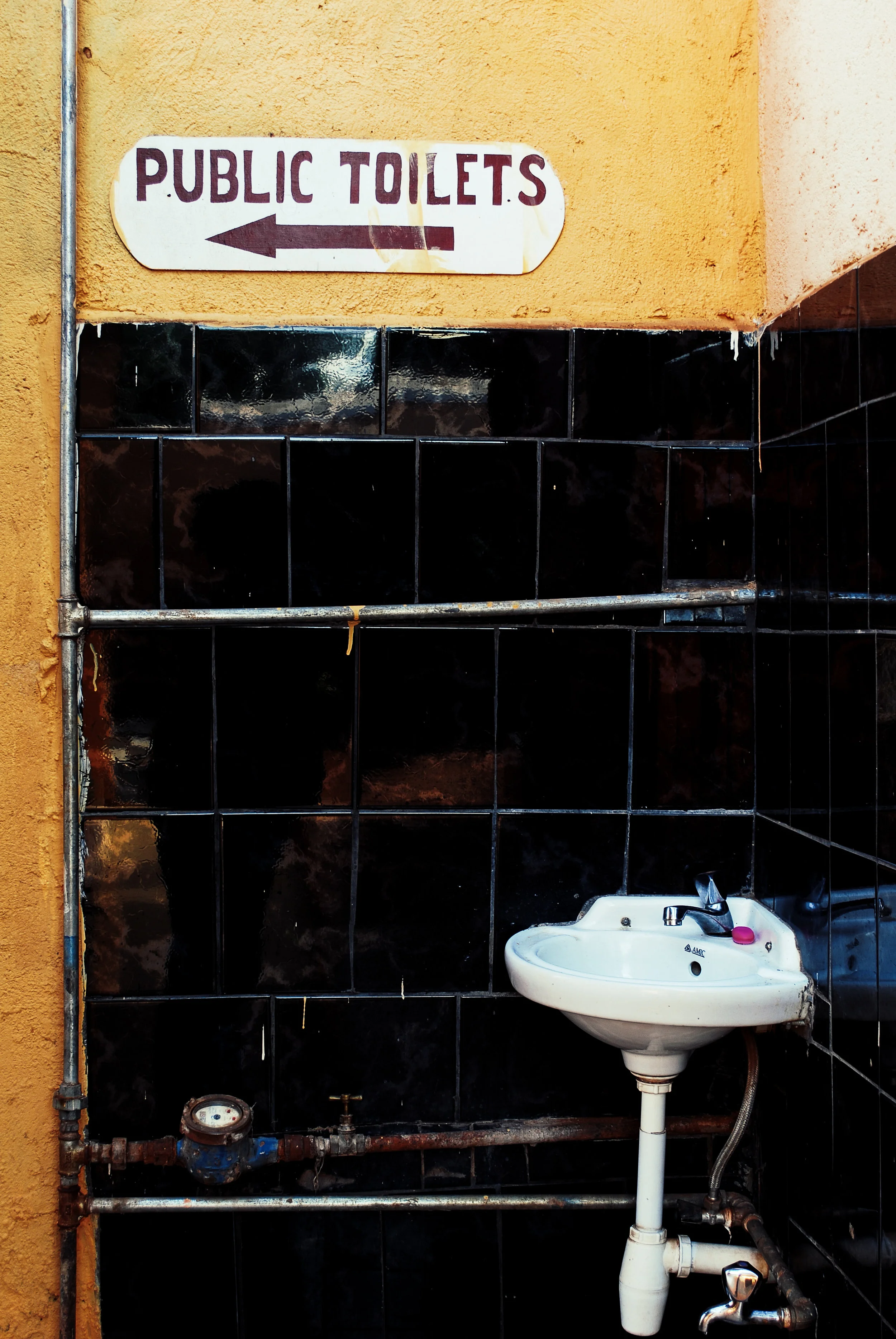 A public restroom with a small sink and plumbing pipes. There is a sign above that points to the left indicating the direction to the public toilets in Entebbe, Uganda.
