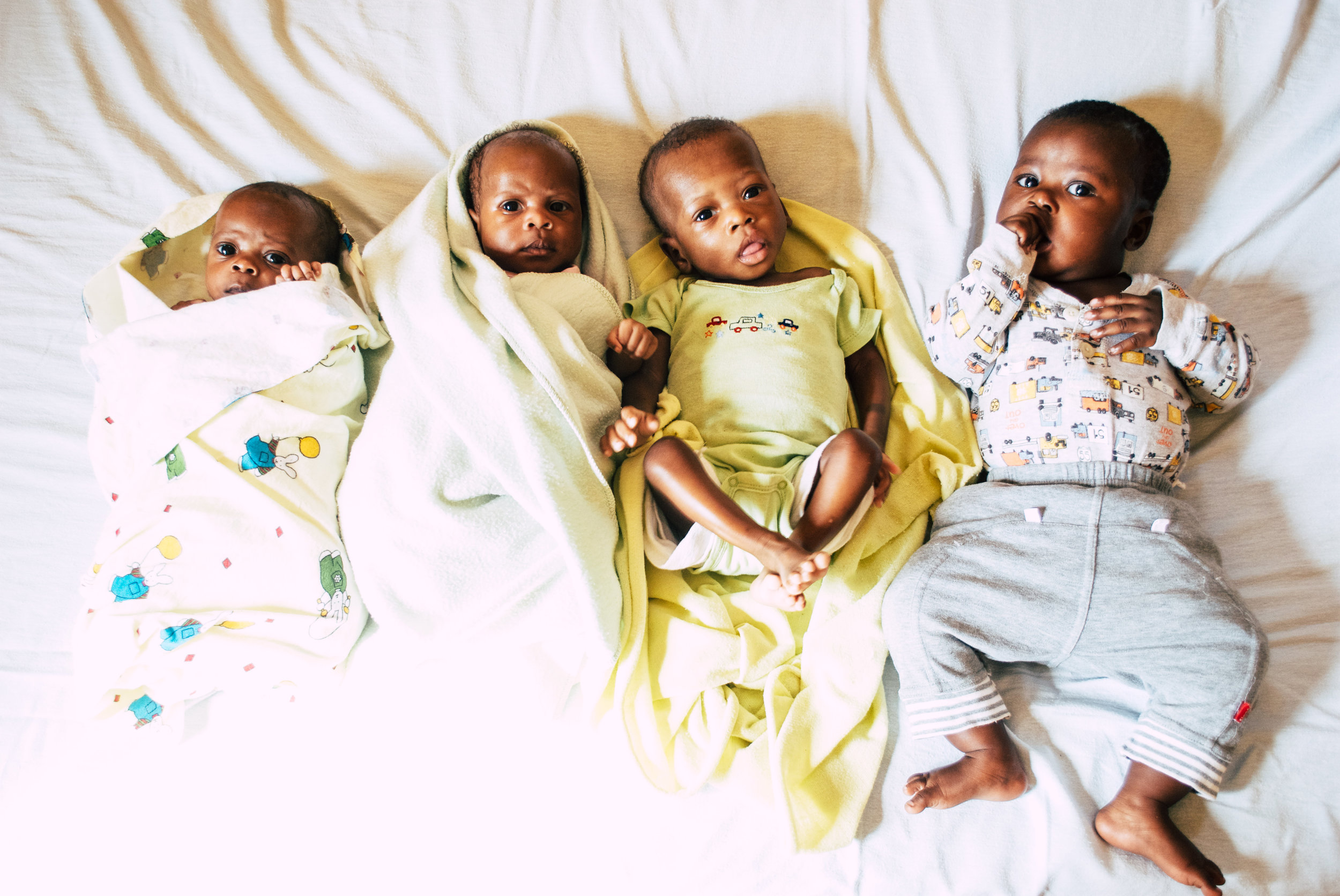 Four young children lying on a bed, three wrapped in blankets and one in pajamas, looking at the camera with different expressions in Uganda.