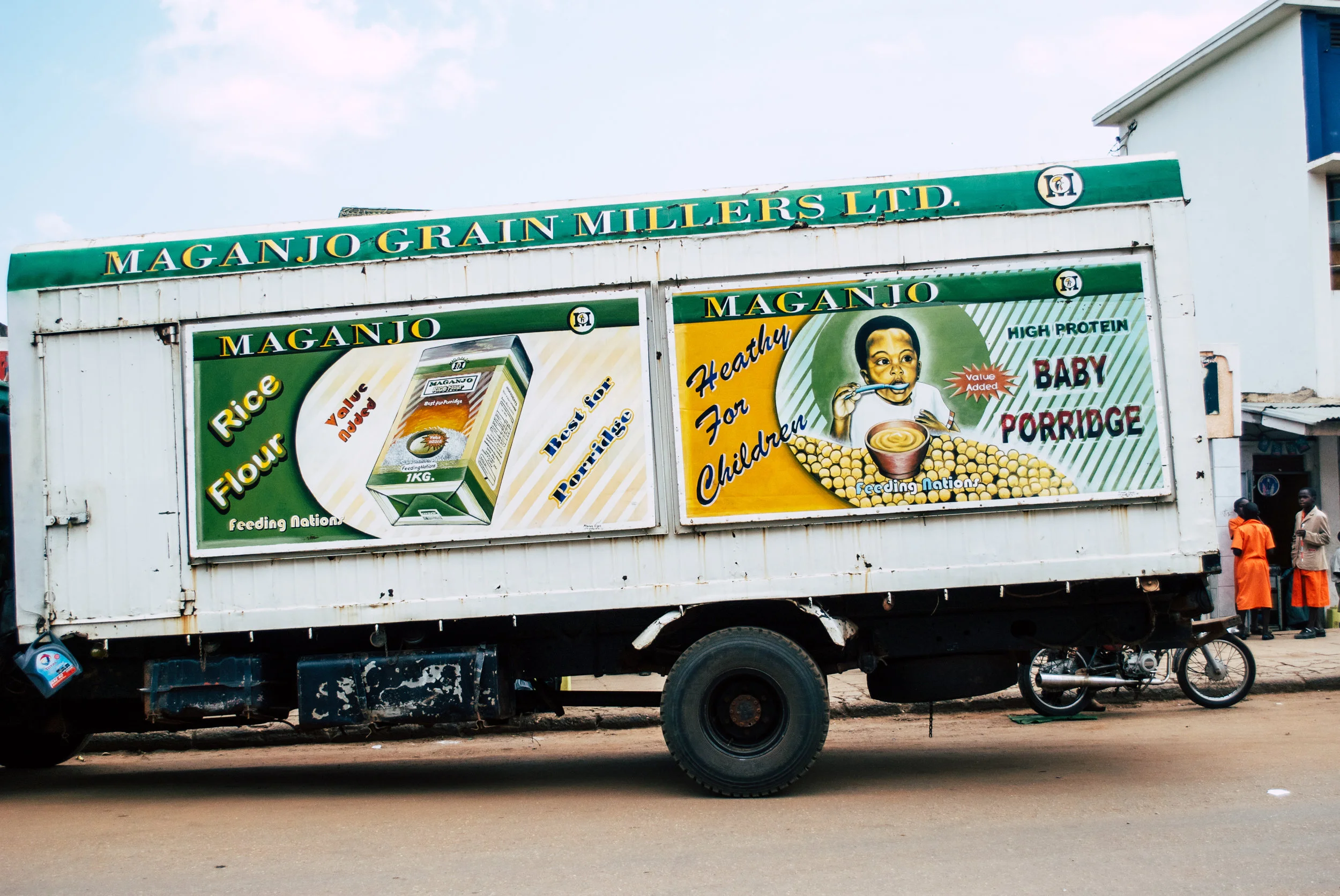 A truck with advertisements for Maganjo Grain Millers Ltd., showing rice flour and baby porridge products, with illustrations of packaging and a child eating porridge, on a street with two women standing nearby in Uganda.