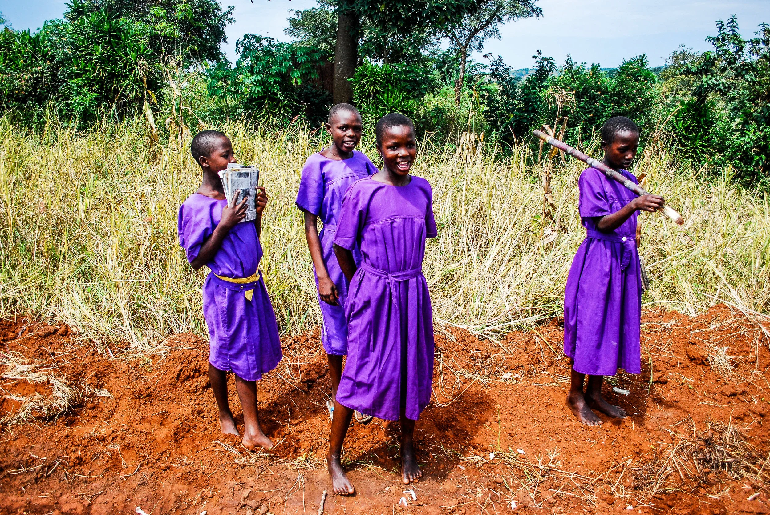 Four young girls in purple dresses stand barefoot on dirt in a field with tall grass, holding farming tools and a bundle of papers, smiling and looking at the camera, with green trees and blue sky in the background in Uganda, East Africa.
