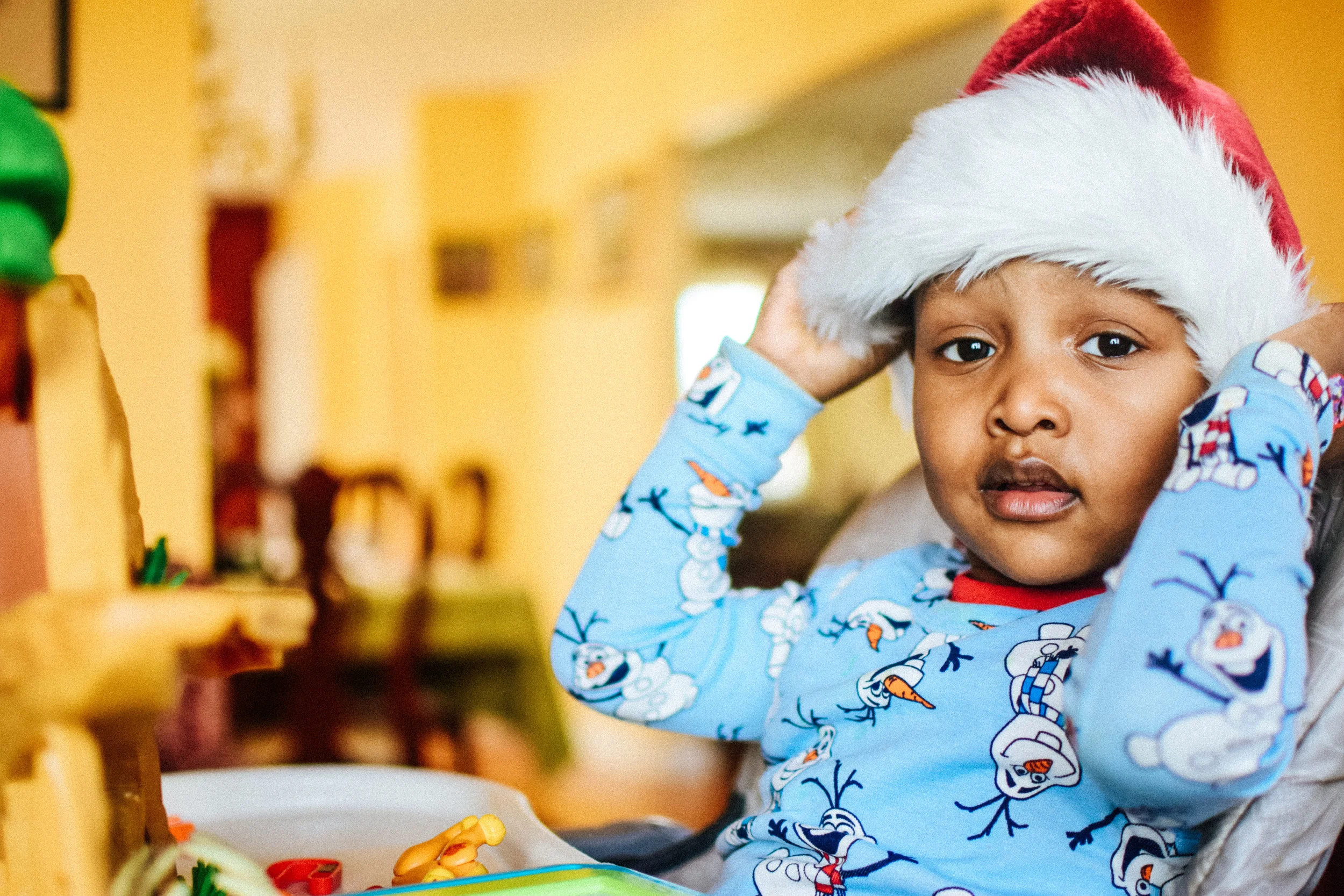 A young child wearing a blue Olaf pajamas and a red Santa hat adjusts the hat on their head, sitting in a home environment.