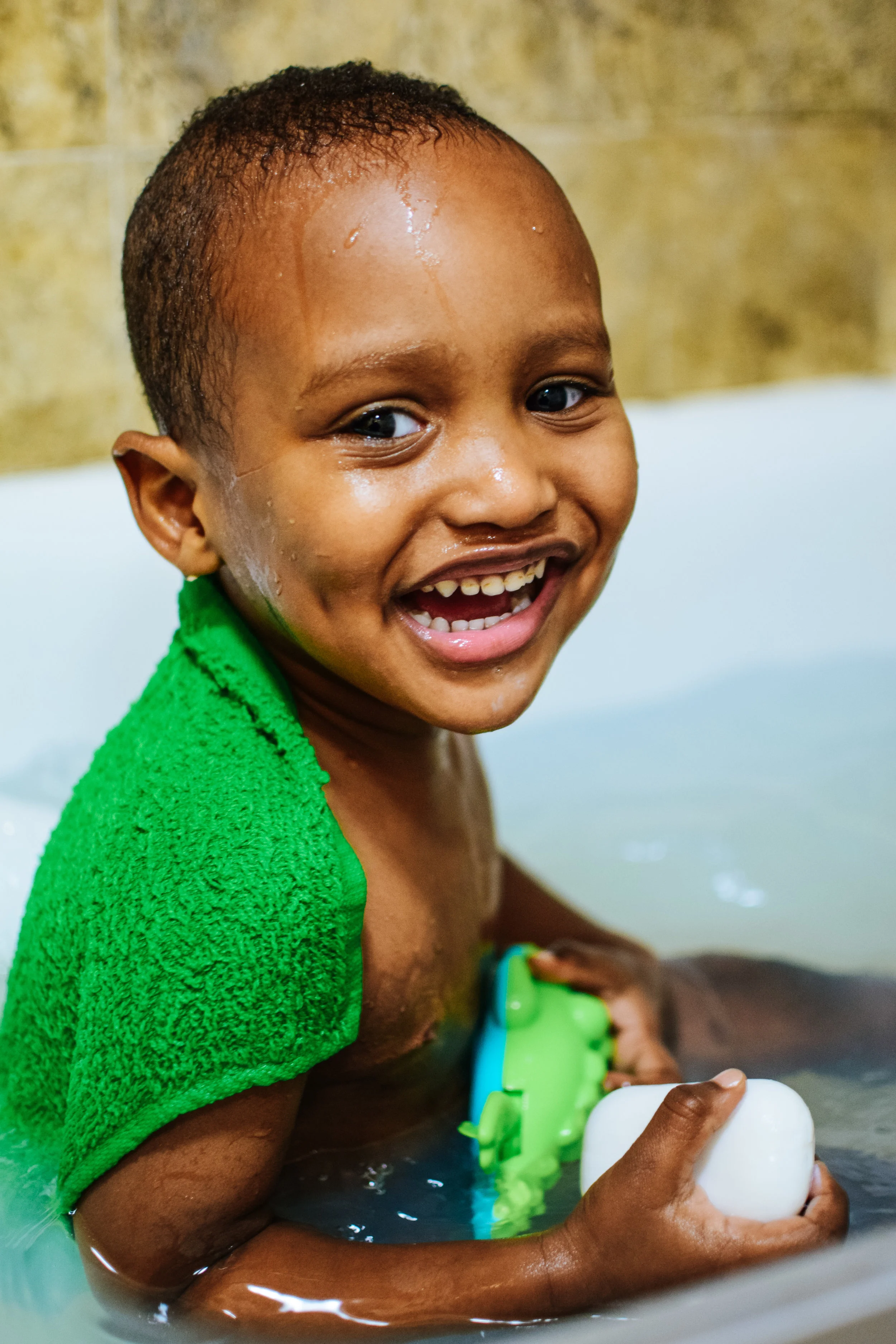 A smiling young boy with short curly hair, wearing a green towel on his shoulders, bathing in a tub with water and holding a bar of soap.