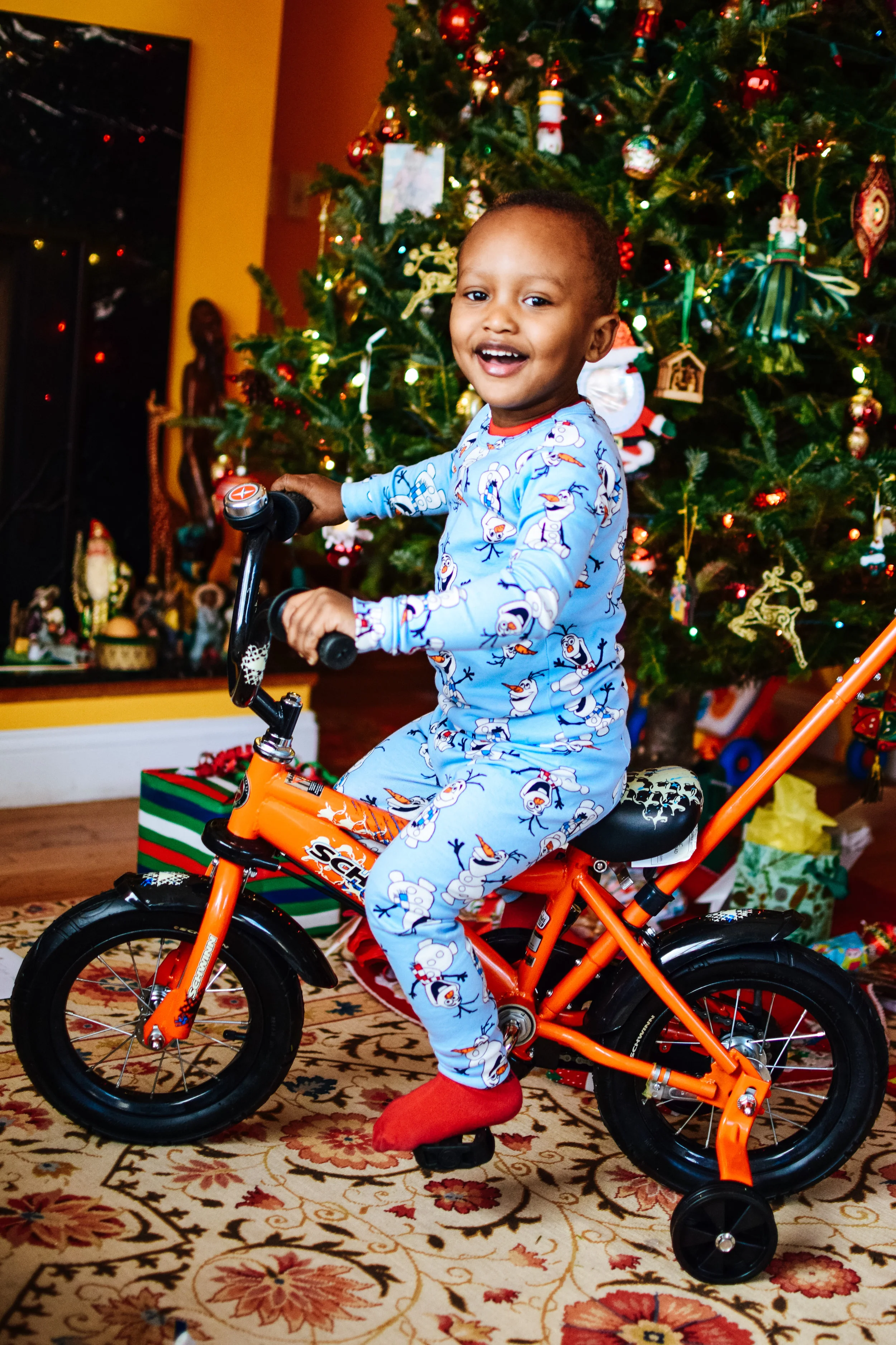 A young boy in Olaf Olafsson pajamas riding a small orange bicycle with training wheels in a living room decorated for Christmas, with a decorated Christmas tree and presents underneath.