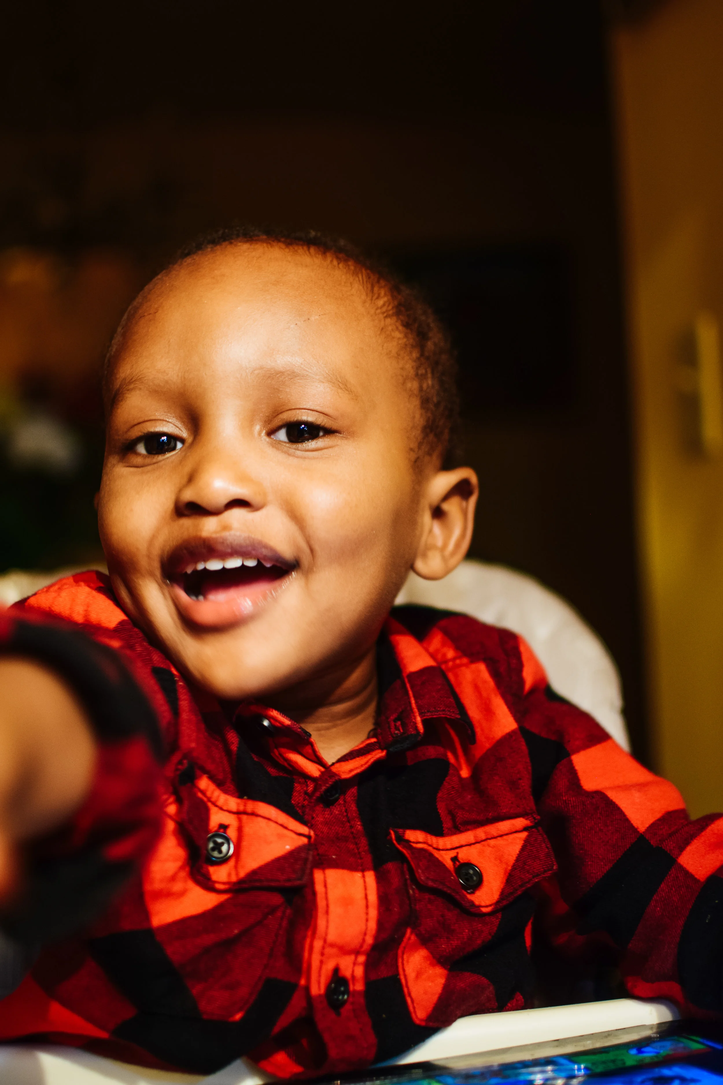 A smiling young boy with short hair wearing a red and black plaid shirt, reaching toward the camera.