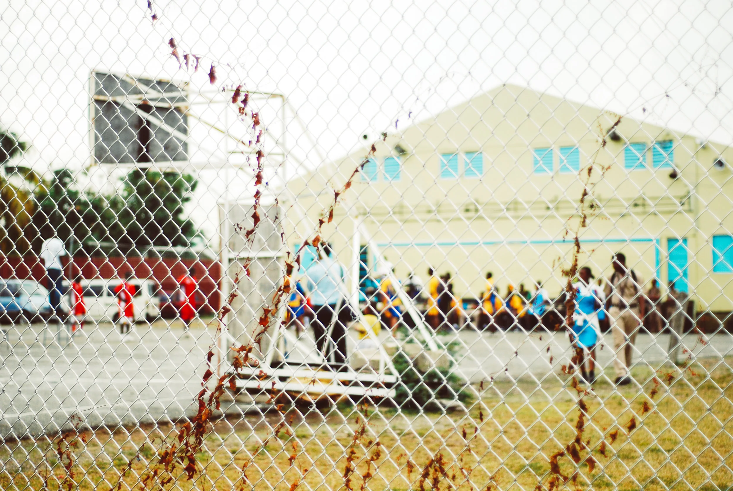 A school playground seen through a chain-link fence, with students in uniforms playing and walking near a yellow building in the background.
