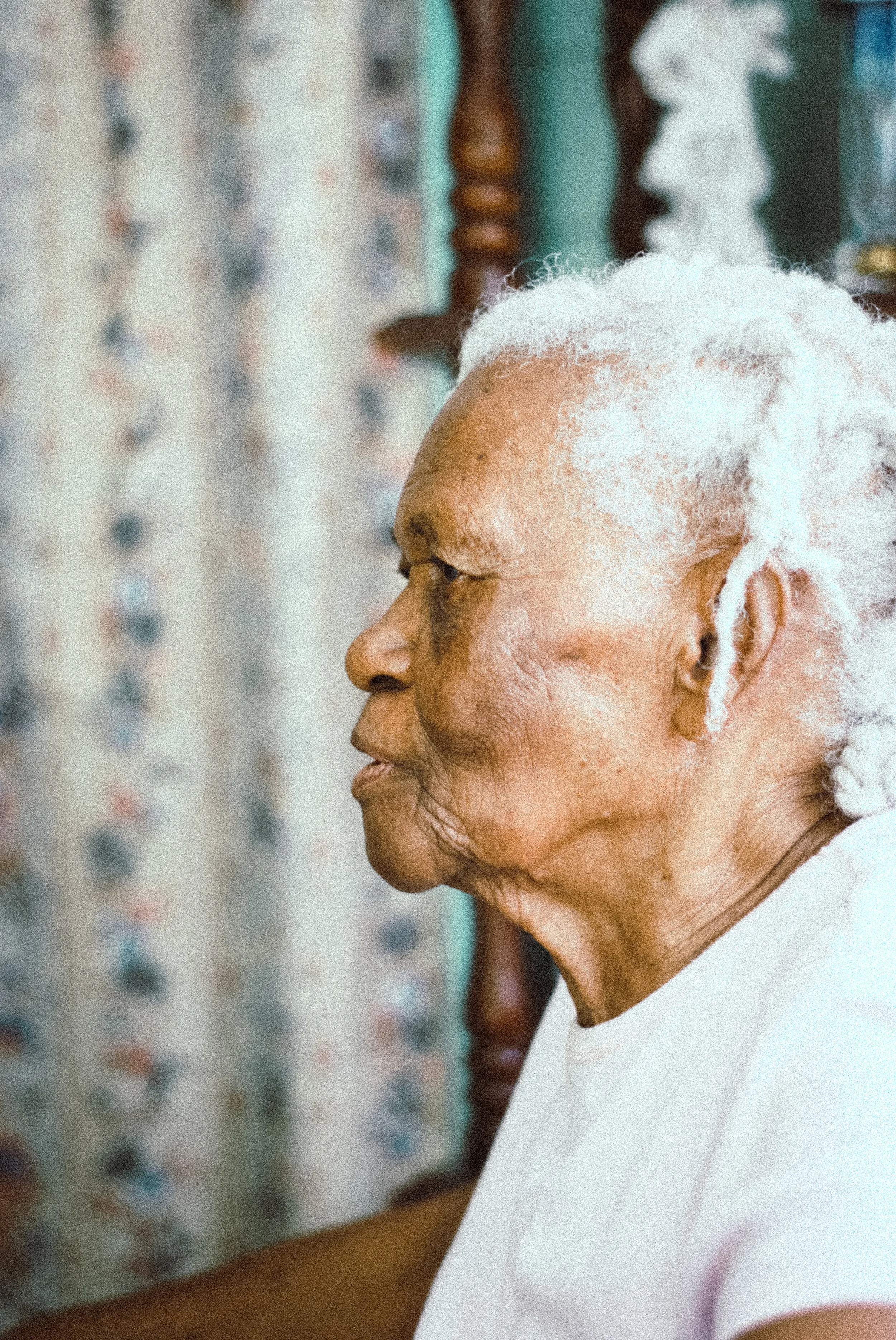 A profile view of an elderly woman with white dreadlocks, wearing a white shirt, sitting indoors with a patterned curtain in the background.